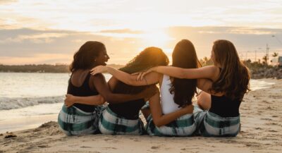 a group of four women with their backs to the camera arms around each other, sit on the beach watching the sunset in the distance.