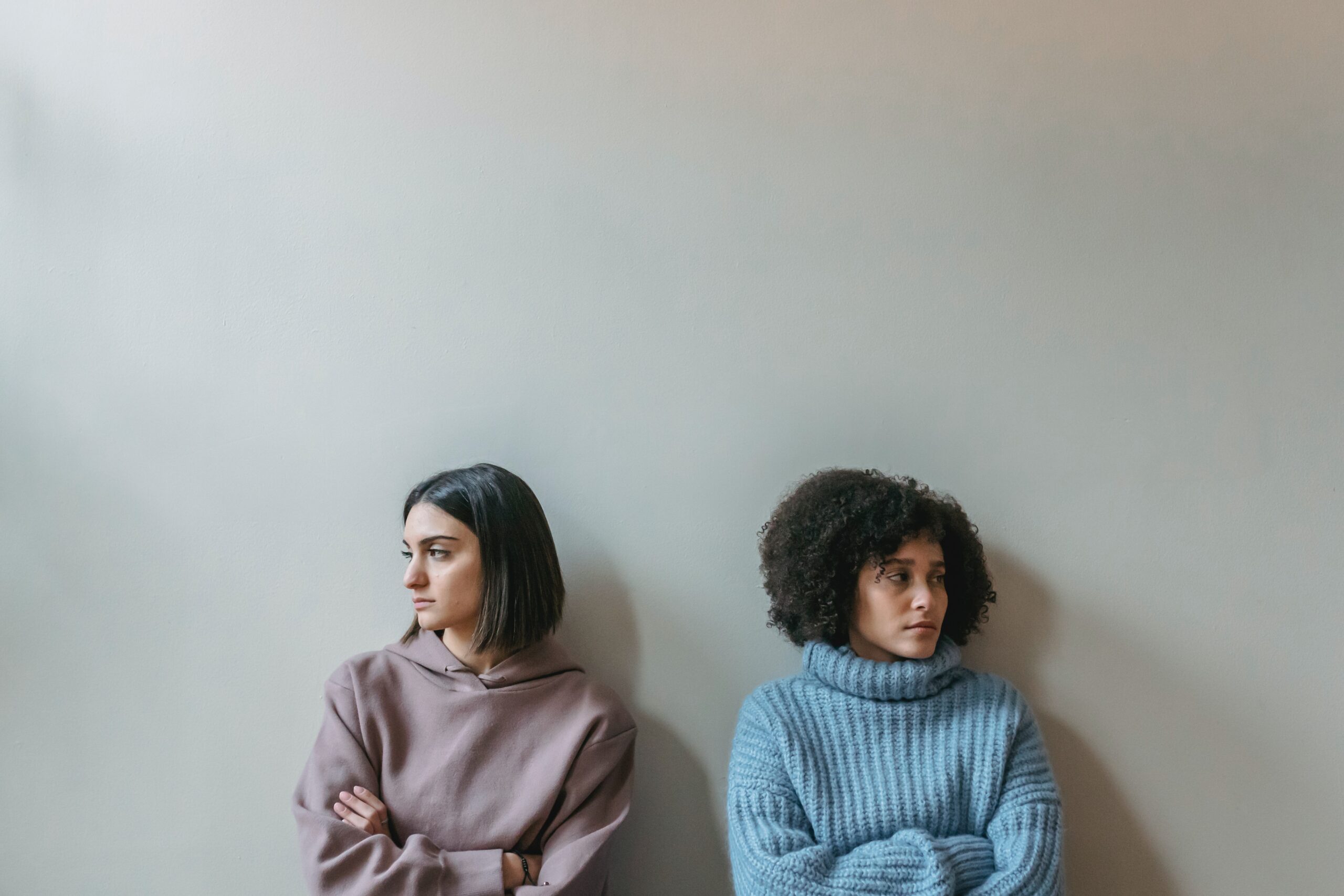 two young women stand against a wall their arms crossed at their chest facing away from one another.